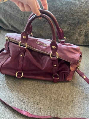 Lila handväska från Miu Miu - Säljer en lila handväska från Miu Miu i mjukt läder med guldfärgade metalldetaljer. Väskan har dubbla handtag, avtagbar axelrem och dragkedja upptill. Klassisk och rymlig modell med snygga ringdetaljer och logga framtill.