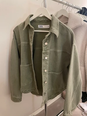 Grön jeansjacka från Zara - Snygg olivgrön jeansjacka från Zara storlek S med raka linjer och två stora fickor framtill. Klassisk krage och silverfärgade knappar. Perfekt oversized passform för en avslappnad look. Tillverkad i jeansmaterial.