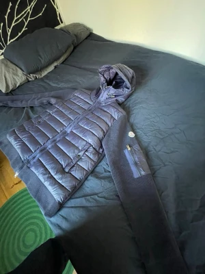 Mörkblå dunväst med huva från Moncler - Snygg mörkblå dunväst från Moncler med stickade ärmar och huva. Västen har dragkedja framtill, fickor med knappar och en extra ficka på ärmen. Insidan har ett unikt serietidningsmönster. Perfekt för dig som vill ha något både stilrent och bekvämt.