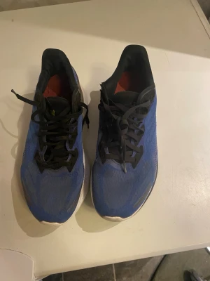 Blå sneakers med svarta detaljer - Snygga blå sneakers med svarta snören och detaljer, samt vit och grön sula. Ovandelen är i meshmaterial för bra andningsförmåga och komfort. Perfekta för dig som gillar sportig stil och vill ha ett par lätta skor till vardagslooken. Har en liten repa i tyget