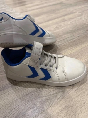 Vita och blå sneakers från Hummel - Snygga vita sneakers från Hummel med blå detaljer och klassiska pilar på sidan. Skorna har både snörning och kardborreband för extra bra passform. Insidan är fodrad i blått tyg och ovandelen är i slätt syntetmaterial.