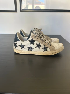 Zadig & Voltaire sneakers med stjärnor - Snygga vita sneakers från Zadig & Voltaire med blåa stjärnor på sidorna och matchande blåa detaljer. Skorna har vita snören med logotyptext och en klassisk låg siluett. 100% läder