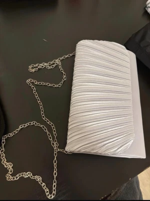 Silver kuvertväska med kedja - Snygg silverfärgad kuvertväska med veckad framsida och elegant metallkedja som axelrem. Väskan har en stilren, rektangulär form och är tillverkad i ett glansigt material som ger en lyxig känsla. Perfekt accessoar för att lyfta din outfit.