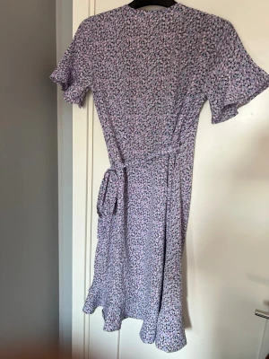 Ljuslila blommig omlottklänning - Söt ljuslila omlottklänning med småblommigt mönster i vitt, grönt och rosa. Klänningen har korta ärmar, volangkant nertill och knytband i midjan. Perfekt för varma dagar och ger en romantisk vibe.