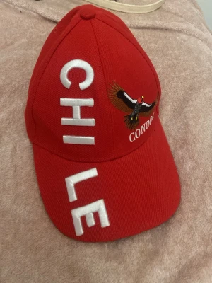 Röd keps med CHILE-tryck och kondor - Snygg röd keps med texten CHILE i stora vita bokstäver och en broderad kondor på sidan. Kepsen har böjd skärm och justerbar baksida. Materialet är troligtvis bomull eller bomullsblandning. Perfekt för dig som vill sticka ut och visa kärlek till Chile.