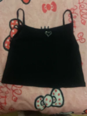 Svart croppad linnetopp med hjärtdetalj - Snygg svart croppad linnetopp med tunna axelband och en liten glittrig hjärtdetalj framtill. Perfekt för dig som gillar enkel men söt stil. Passar till både jeans och kjol.