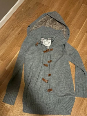 Grå kofta med luva och träknappar - Mysig grå stickad kofta med stor luva fodrad i mjuk fuskpäls. Koftan har långa ärmar, ribbade muddar och stängs med snygga träknappar. Mennan M o L i storlek 