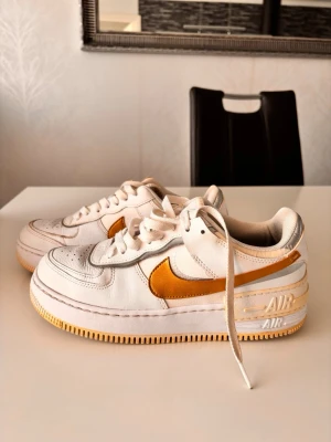 Nike Air Force 1 vita och guld sneakers - Nike Air Force 1 sneakers i vitt läder med guld swoosh och silver detaljer. Klassisk låg modell med snörning och AIR-tryck på sulan. Yttersulan är ljusgul och insidan är ljusbeige. Perfekt för dig som gillar retrostil och streetwear.