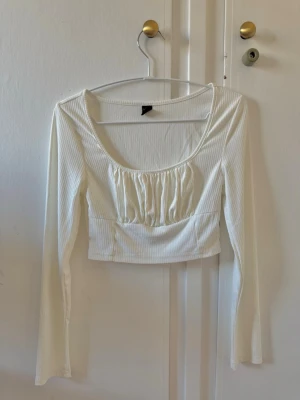 Vit ribbad croppad topp - Säljer en vit croppad topp med långa ärmar och ribbad struktur. Toppen har en djup rundad urringning och rynkade detaljer framtill som ger en snygg form. Perfekt att styla med höga jeans eller kjol.