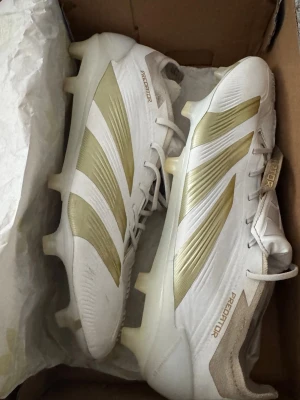 Adidas Predator vita/guld fotbollsskor - Säljer ett par Adidas Predator fotbollsskor i vitt med guldiga detaljer och klassiska tre ränder på sidan. Skorna har snörning, mönstrad ovandel och fasta dobbar för spel på gräs. Tillverkade i syntetmaterial för bra grepp och kontroll på planen.                                                               Observera att byte till en annan skomodell i samma storlek också är möjligt.