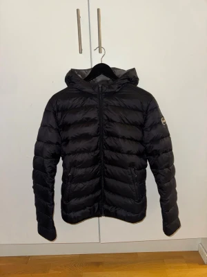 Colmar jacka dam - Snygg svart dunjacka med huva och dragkedja framtill. Jackan har två sidofickor med dragkedja och quiltad design. Märke syns på vänster ärm. Perfekt för kalla vinterdagar och ger en sportig look.