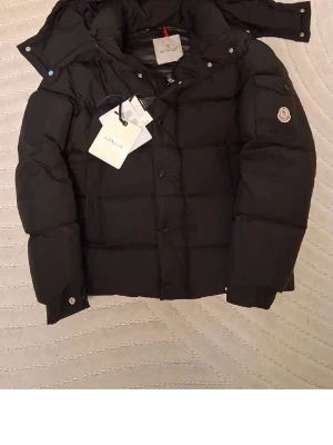 Svart dunjacka från Moncler - Säljer en svart dunjacka från Moncler med avtagbar huva och klassisk Moncler-logga på ärmen. Jackan har stora, vadderade paneler, fickor med lock och tryckknappar samt dragkedja och knappstängning framtill. Perfekt för kalla vinterdagar.