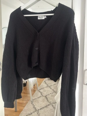 Svart ribbad kofta från NA-KD - Säljer en svart ribbstickad kofta från NA-KD med v-ringning och knäppning framtill. Koftan har långa, lite pösiga ärmar och en croppad passform.