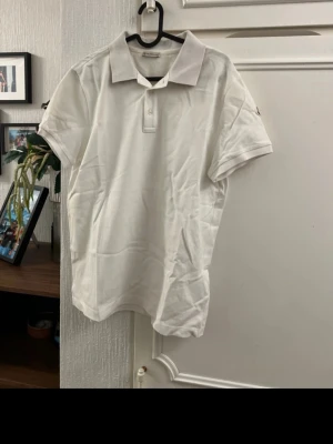 Vit pikétröja från Moncler - Klassisk vit pikétröja från Moncler med krage och knappar framtill. Tillverkad i bomull för en bekväm känsla och stilren look. Perfekt till jeans eller shorts för en enkel men snygg outfit.