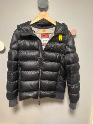 Parajumper jacka  - Snygg svart dunjacka från Parajumpers med huva, gul detalj vid halsen och patch på ärmen. Jackan har breda, quiltade paneler, dragkedja framtill och muddar vid ärmsluten. Perfekt för kalla vinterdagar och riktigt trendig look. Köpte den av min kusin som köpte den fån nk Stockholm! Skannar man qr koden kommer man direkt till pj’s hemsida!