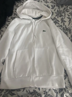 Vit hoodie med dragkedja från Lacoste - Säljer en stilren vit hoodie från Lacoste med dragkedja framtill och två stora fickor. Klassisk huva med snörning och den ikoniska gröna krokodilloggan på bröstet. Perfekt för en avslappnad look och enkel att matcha med det mesta.
