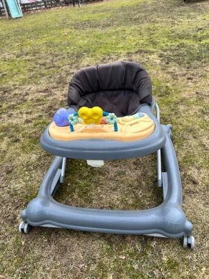 Grå gåstol med leksaker - En grå gåstol med mjuk svart sits och lekbricka i gult och blått. Lekbrickan har färgglada detaljer och roliga figurer för barnet att leka med. Stolen har fyra hjul och en stabil ram i plast och metall. Perfekt för småbarn som vill utforska och röra sig mer.