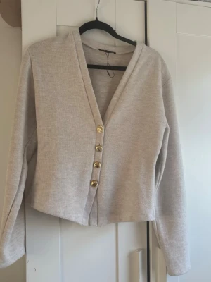 Kofta med knappar - Stilren beige kofta med v-ringning och fyra guldfärgade knappar framtill. Koftan har lång ärm och är tillverkad i ett mjukt, stickat material. Storlek M men passar också som S