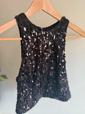 Svart paljettopp med rund hals och dragkedja  - Snygg svart ärmlös topp täckt med glittriga paljetter som reflekterar ljuset. Modellen är croppad med rund halsringning och har en festlig vibe. Perfekt att för att styla en cool look eller till festival. Dragkedja bak. Använd en gång.