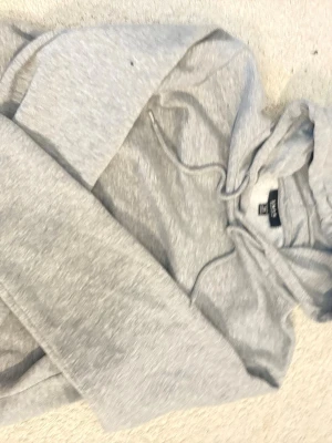 Ljusgrå hoodie från SMOG - En stilren ljusgrå hoodie från SMOG i storlek XS. Hoodien har en klassisk huva med dragsko och är tillverkad i mjukt material med borstad insida för extra komfort. Helt fläckfri men ganska använd. Perfekt för en avslappnad look och enkel att matcha med andra plagg.