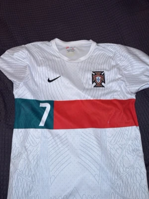 Portugal Nike fotbollströja nr 7 - Säljer en vit Portugal fotbollströja från Nike med nummer 7 på bröstet. Tröjan har grönt och rött band över magen, broderad Portugal-emblem och Nike-logga. Tillverkad i Dri-FIT material som andas, perfekt för träning eller match.