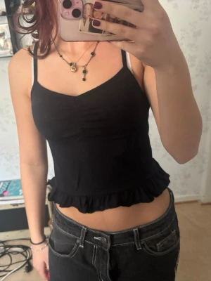 Svart croppad linnetopp med volang - Säljer en svart croppad linnetopp med smala axelband och volangkant nedtill. Toppen har en lätt v-ringad urringning och är perfekt för dig som gillar en enkel men söt stil. Materialet känns mjukt och stretchigt.