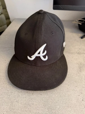 Svart New Era keps med A-logga - Snygg svart keps från New Era med vit broderad A-logga framtill och New Era-logga på sidan. Klassisk rak skärm och strukturerad passform. Perfekt för dig som gillar streetwear och sportig stil.