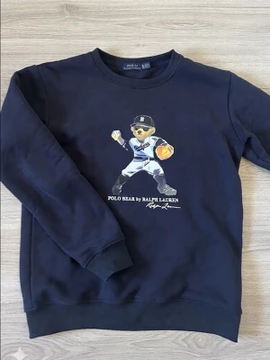 Svart sweatshirt Polo Ralph Lauren XL - Svart sweatshirt från Polo Ralph Lauren med ikoniskt Polo Bear-motiv på bröstet. Rund halsringning och ribbade muddar. Perfekt för dig som gillar streetwear och vill ha en klassisk men lekfull look.