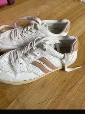 Vita sneakers med beige detaljer - Snygga vita sneakers med perforerat mönster och två beige ränder på sidan. Skorna har vita snören och beige detalj på hälen. 