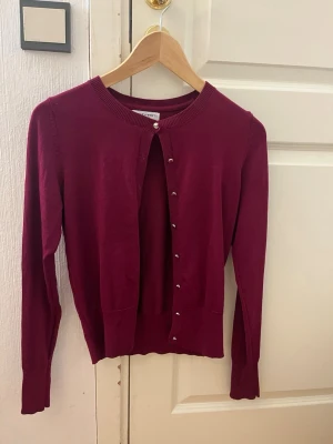 Vinröd kofta från Holly & Whyte - Snygg vinröd kofta från Holly & Whyte by Lindex. Klassisk rund halsringning, ribbade muddar och silverfärgade knappar framtill. Perfekt att slänga över en topp eller klänning. Tillverkad i mjukt stickat material.