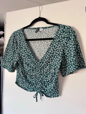 Grön/vit blommig topp med snörning fram - Supersöt grön topp från H&M med vitt blommönster. Perfekt för vår och sommar när du vill ha något lätt och fräscht. Sparsamt använd.