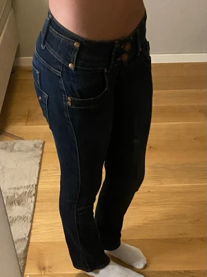 Mörkblå jeans med låg midja - Snygga mörkblå jeans med låg midja och raka ben. Jeansen har dubbla knappar framtill, klassiska fem fickor och dekorativa sömmar. Materialet är denim med stretch för skön passform. Perfekta till en avslappnad stil.