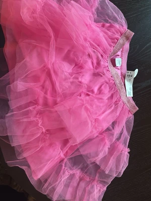 Rosa tyllkjol från Lindex - Supersöt rosa tyllkjol från Lindex med flera lager volang och ett glittrigt resårband i midjan. Kjolen har en fluffig look och passar perfekt för dig som vill sticka ut med färg och volym.