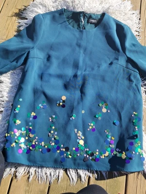 Blå paljett t-shirt Lawrence Grey - Säljer en blå t shirt från Lawrence Grey med korta ärmar och rund hals. T shirten har stora, färgglada paljetter längs nederkanten som ger en unik och festlig look. 