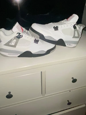 Air Jordan 4 White Cement  - Säljer ett par Air Jordan 4 White Cement sneakers med vit ovandel i läder, grå och svarta detaljer samt meshpaneler på sidorna. Skorna har den klassiska Jumpman-loggan på plösen och synlig Air-enhet i sulan. Perfekta för dig som gillar retrostil och streetwear.
