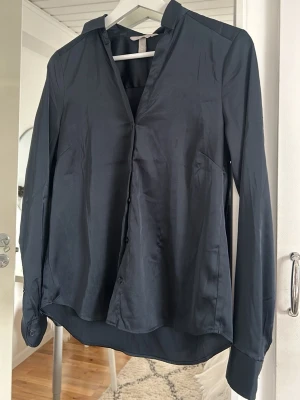 Mörkblå satinblus från H&M - Stilren svart blus i glansigt satinmaterial från H&M. Klassisk krage, knäppning framtill och långa ärmar med knapp vid ärmslut. Super skönt material 
