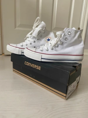 Vita Converse Chuck Taylor All Star - Helt nya Klassiska vita Converse Chuck Taylor All Star high tops med vit canvas, vita snören och gummisula. Skorna har den ikoniska loggan på sidan och röda samt blå detaljer runt sulan. Perfekta för en avslappnad och stilren look.