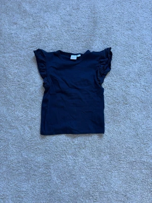 Marinblå topp med volangärmar - En marinblå topp med volangdetaljer på ärmarna. Modellen är ärmlös med breda axelpartier och har en enkel, rak passform. Perfekt att styla med jeans eller kjol för en trendig look.