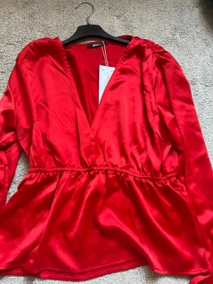 Röd satinblus med v-ringning - Snygg röd blus från Ginatricot i glansigt satintyg. Blusen har djup v-ringning, markerad midja med resår och långa ärmar. Perfekt för dig som vill sticka ut med en färgstark och trendig look.