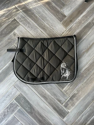 Svart schabrak med hästmotiv - Jättefint full allround schabrak!💓 Säljer ett svart, vadderat schabrak med rutmönster och vit kant. Ena sidan har ett vitt tryck av en häst. Perfekt till ridning och passar under sadeln. Fästen med kardborreband för enkel användning.