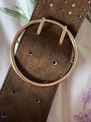 Brett brunt mockabälte med rund spänne - Snyggt och brett bälte i brun mocka från Accessories. Bältet har ett stort, runt spänne i guldtonad metall som ger en trendig look. Perfekt för att lyfta vilken outfit som helst och passar till både jeans och klänning.