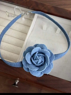 Blått halsband Necklace chocker med tygblomma - Choker-halsband i blått tyg med en stor, detaljrik blomma framtill. Halsbandet har justerbart kedjelås och ger en cool, trendig denimkänsla. Perfekt för att lyfta din outfit med en unik accessoar.