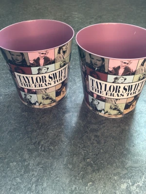 Taylor Swift The Eras Tour popcornhinkar - Två popcornhinkar med Taylor Swift The Eras Tour-tryck. Hinkarna är i metall med rosa insida och collage av olika Taylor Swift-bilder i svartvitt och pastellfärger på utsidan. Perfekt för snacks eller som samlarpryl för fans.