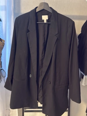 Svart oversized kavaj från H&M - Stilren svart oversized kavaj från H&M med klassisk krage och enkelknäppning. Kavajen har två framfickor och långa ärmar. Perfekt för en trendig och avslappnad look. Materialet är mjukt och följsamt, vilket ger en skön passform.