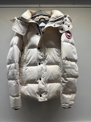 Vit pufferjacka från Canada Goose - Säljer en snygg vit pufferjacka från Canada Goose med ikonisk röd och blå patch på ärmen. Jackan har hög krage, svarta knappar och dragkedja framtill. Fylld med dun och har en clean, boxig siluett som håller dig varm under kalla dagar. Wyndham modell