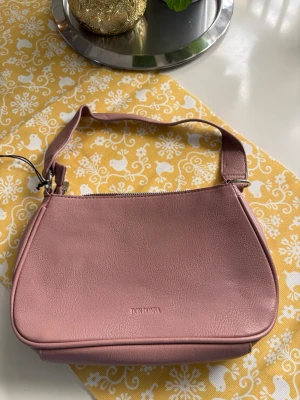 Rosa axelremsväska från Don Donna - Snygg rosa handväska från Don Donna i ett stilrent och mjukt konstläder. Väskan har en enkel axelrem och dragkedja upptill. Perfekt storlek för det viktigaste och en clean look med diskret logga framtill.