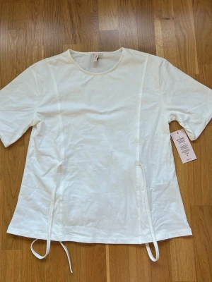 Vit t-shirt med snörning från Nelly - Säljer en vit t-shirt från Nelly i modellen Tie Waist Tee. T-shirten har korta ärmar, rund halsringning och snygga snörningsdetaljer framtill. Tillverkad i mjuk bomull för en skön känsla och enkel stil.