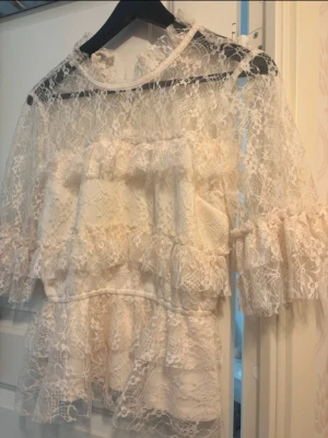 Vit spetsblus med volanger - Romantisk vit blus i genombruten spets med volangdetaljer och hög hals. Blusen har trekvartsärmar och transparenta partier som ger en luftig känsla. Perfekt för dig som gillar en feminin och trendig stil.