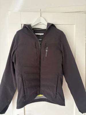 Svart vindjacka från Sail racing  - Snygg svart vindjacka från Sail racing  med huva och dragkedja framtill. Jackan har två sidofickor och en bröstficka med dragkedja samt logga på bröstet. Tillverkad i ett slitstarkt, vattenavvisande material som passar perfekt för blåsiga dagar. 