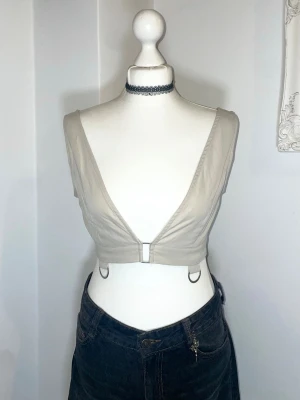 Beige croppad linnetopp med ringdetaljer - Beige/Sten färgad croppad linnetopp med djup v-ringning och silverfärgade ringdetaljer framtill och på sidorna. Toppen är ärmlös och har en stilren, minimalistisk look. UK size 10